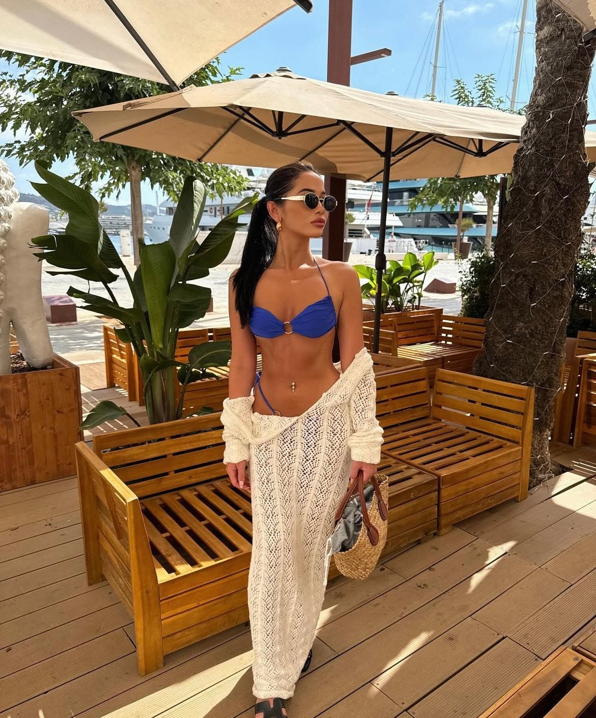Woman in a blue bikini and white cover-up standing on a wooden deck with plants and umbrellas in the background.